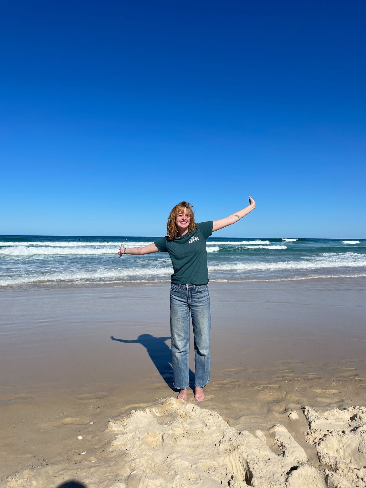 A joyful person with arms outstretched stands on a sandy beach with blue ocean waves and clear sky in the background.