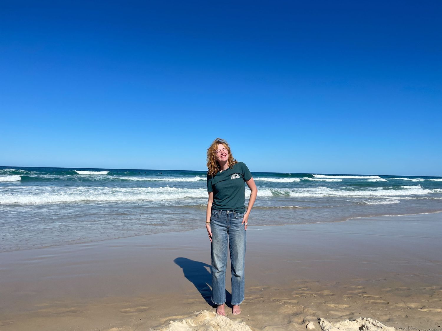 Someone stands on a sunny beach with crashing waves and blue ocean waters under clear skies.