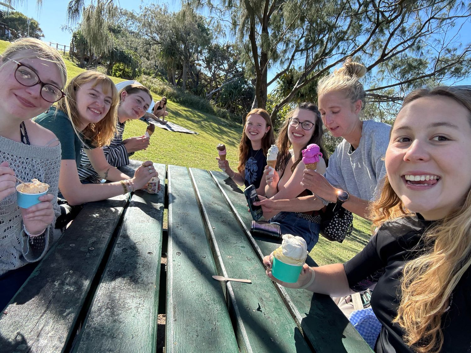 A fun group gathers around outdoor picnic tables sharing food and drinks together on a sunny afternoon.