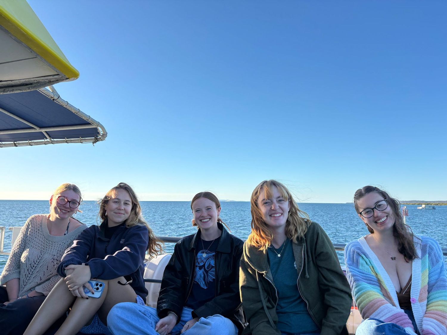 A group of friends enjoying a sunny day together on a boat with blue water and clear skies in the background.