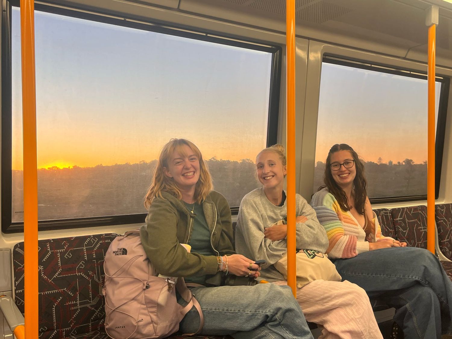Passengers sit together on public transport during sunset with orange sky visible through large windows.