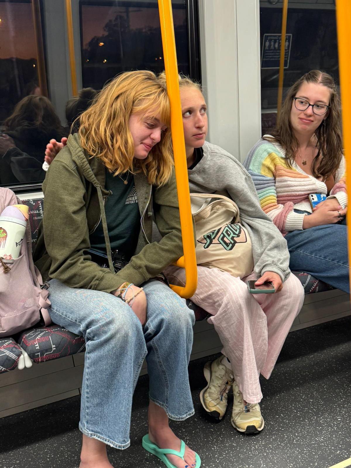 Passengers sit together on public transit seats with one person appearing tired while others look at their phones.