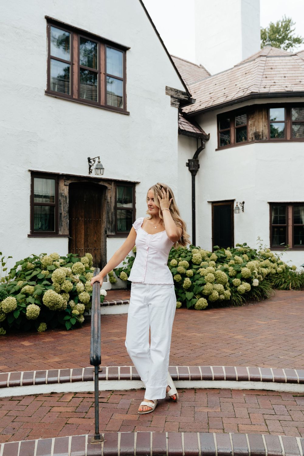 A flowing white dress moves gracefully across brick pavement beside a white building with hydrangeas.