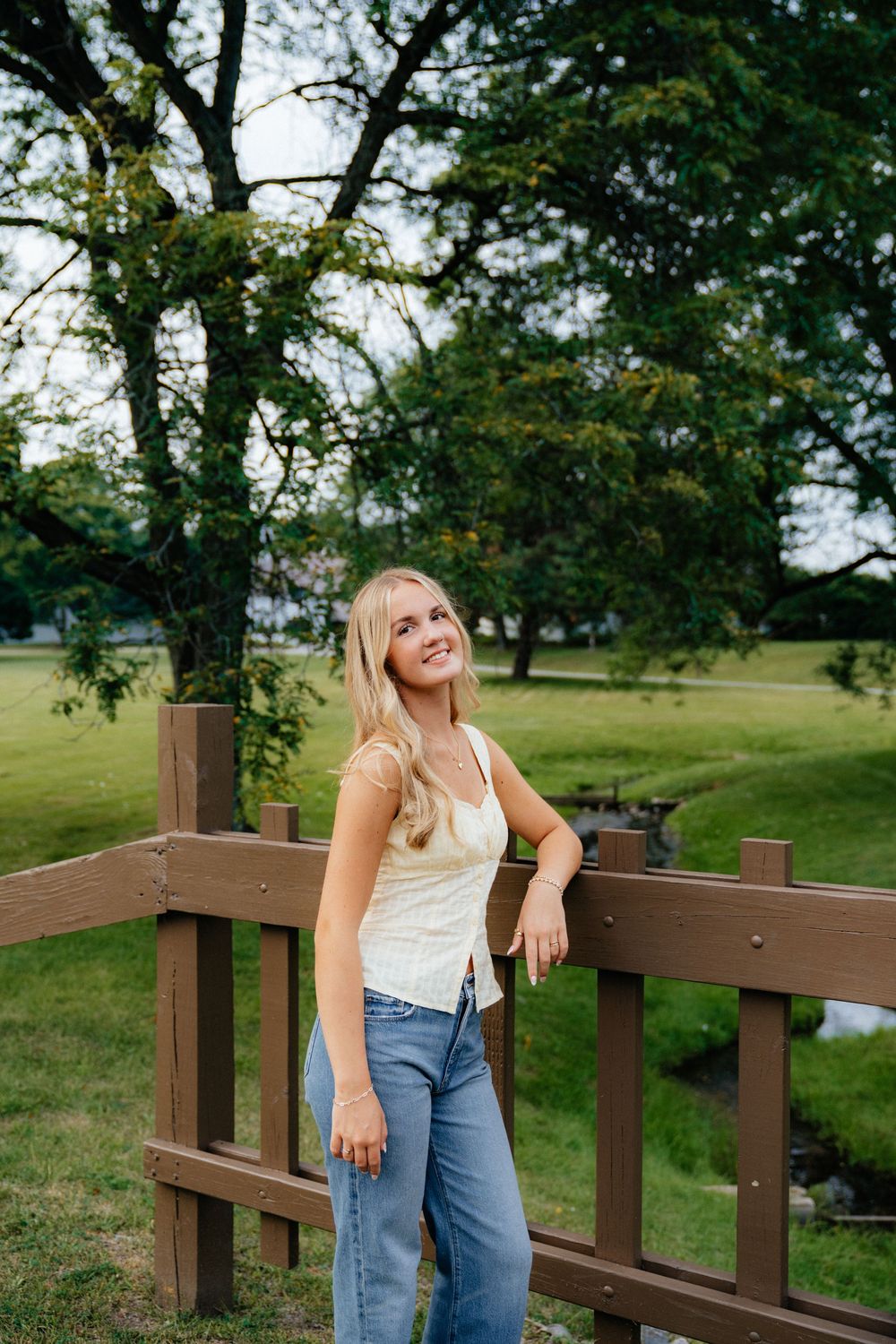A casual summer outfit with jeans and white top while standing on a wooden deck railing in a park setting.