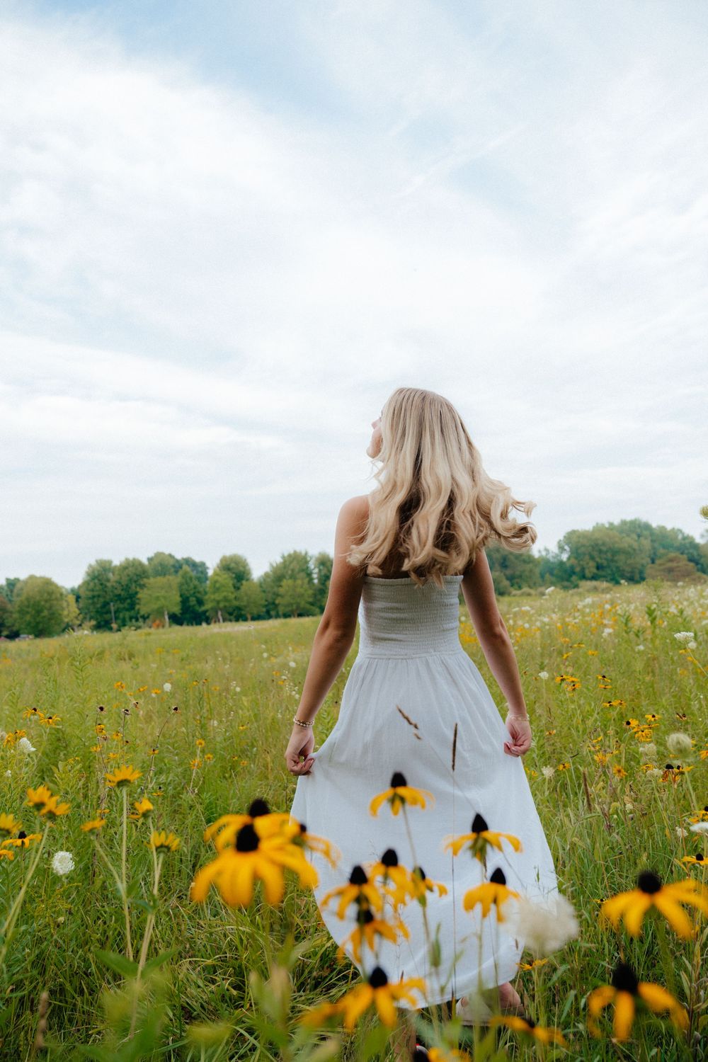 Back view of a flowing white dress in a field of black-eyed susan flowers under cloudy skies.