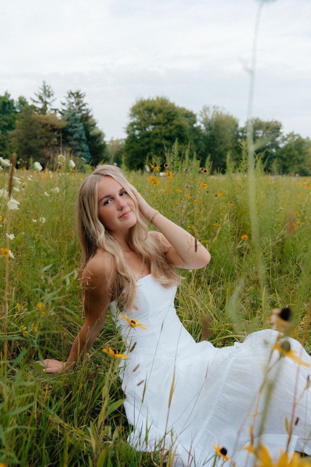 A romantic portrait in a white dress sitting among wildflowers in a summer meadow.