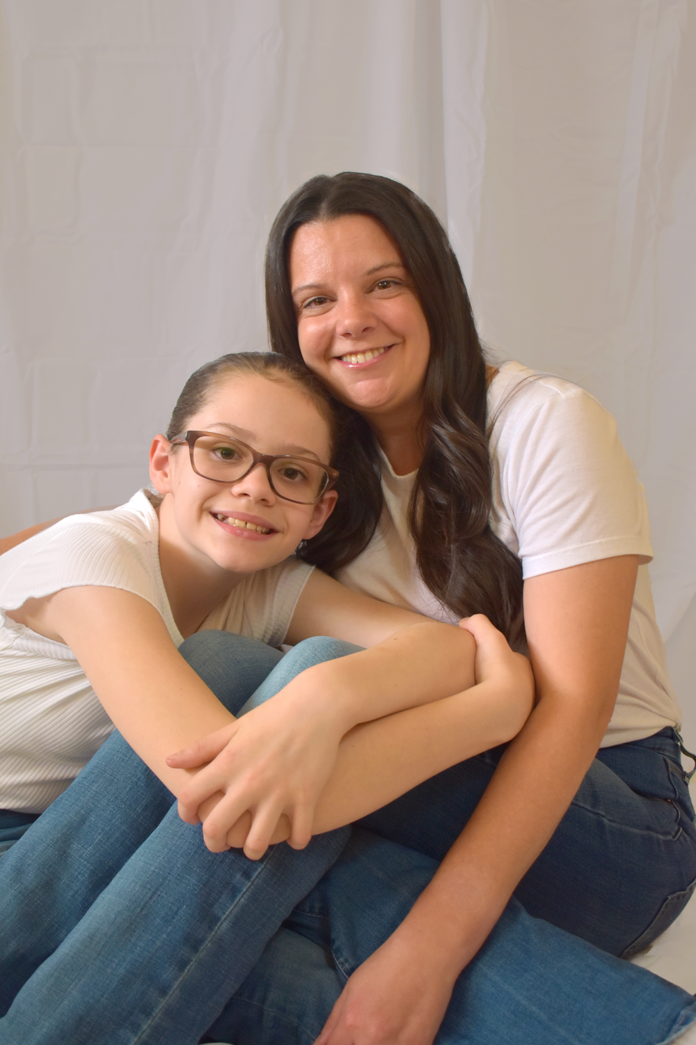 Two people sitting close together on a couch wearing jeans and white shirts, smiling warmly at the camera.