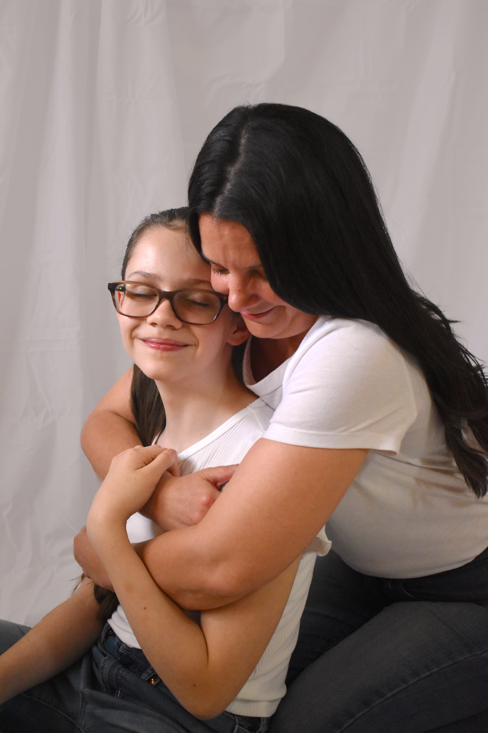 Two people in white t-shirts share an intimate embrace while seated against a light gray backdrop.