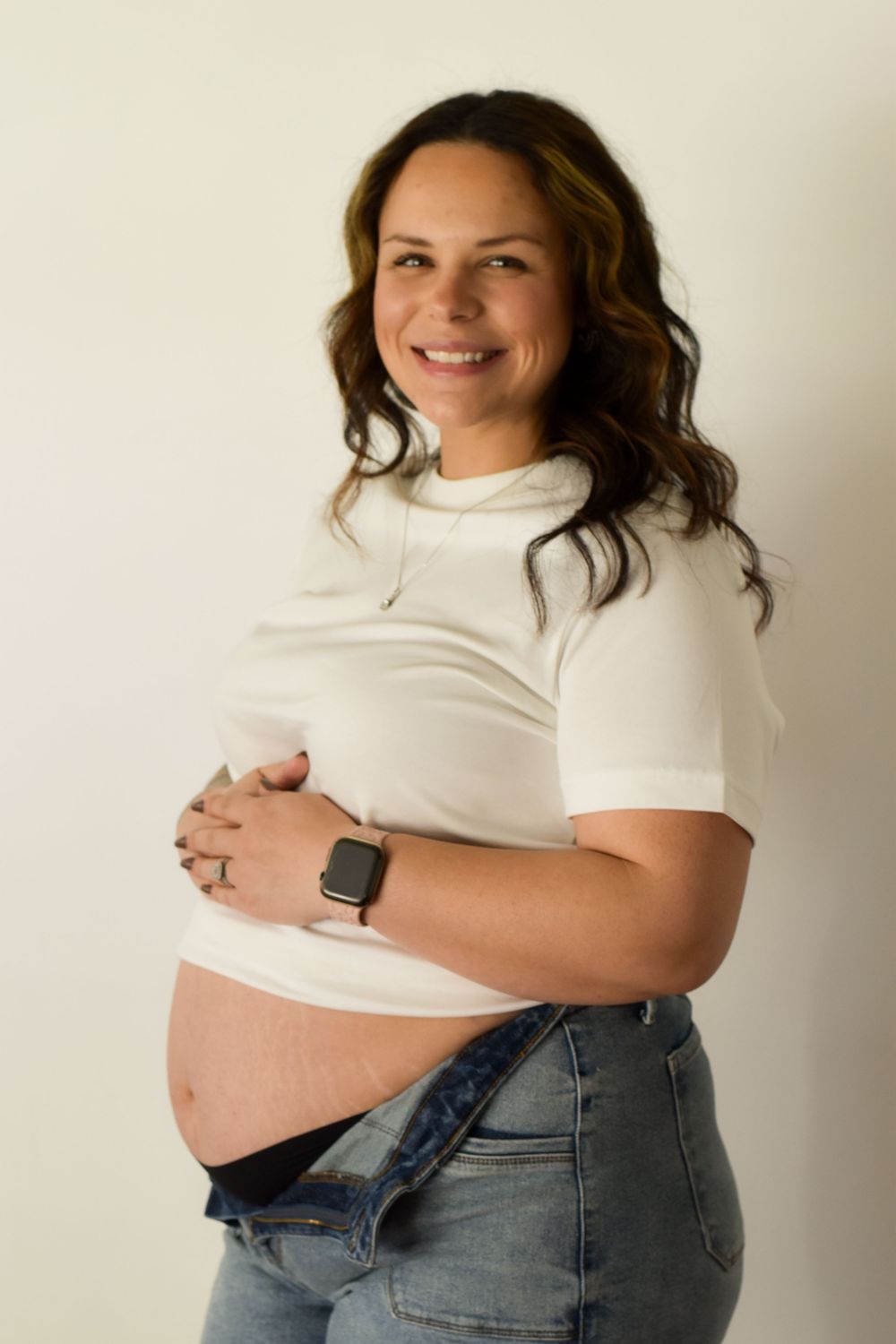 A pregnant woman in white top and jeans stands against white wall showing her baby bump.