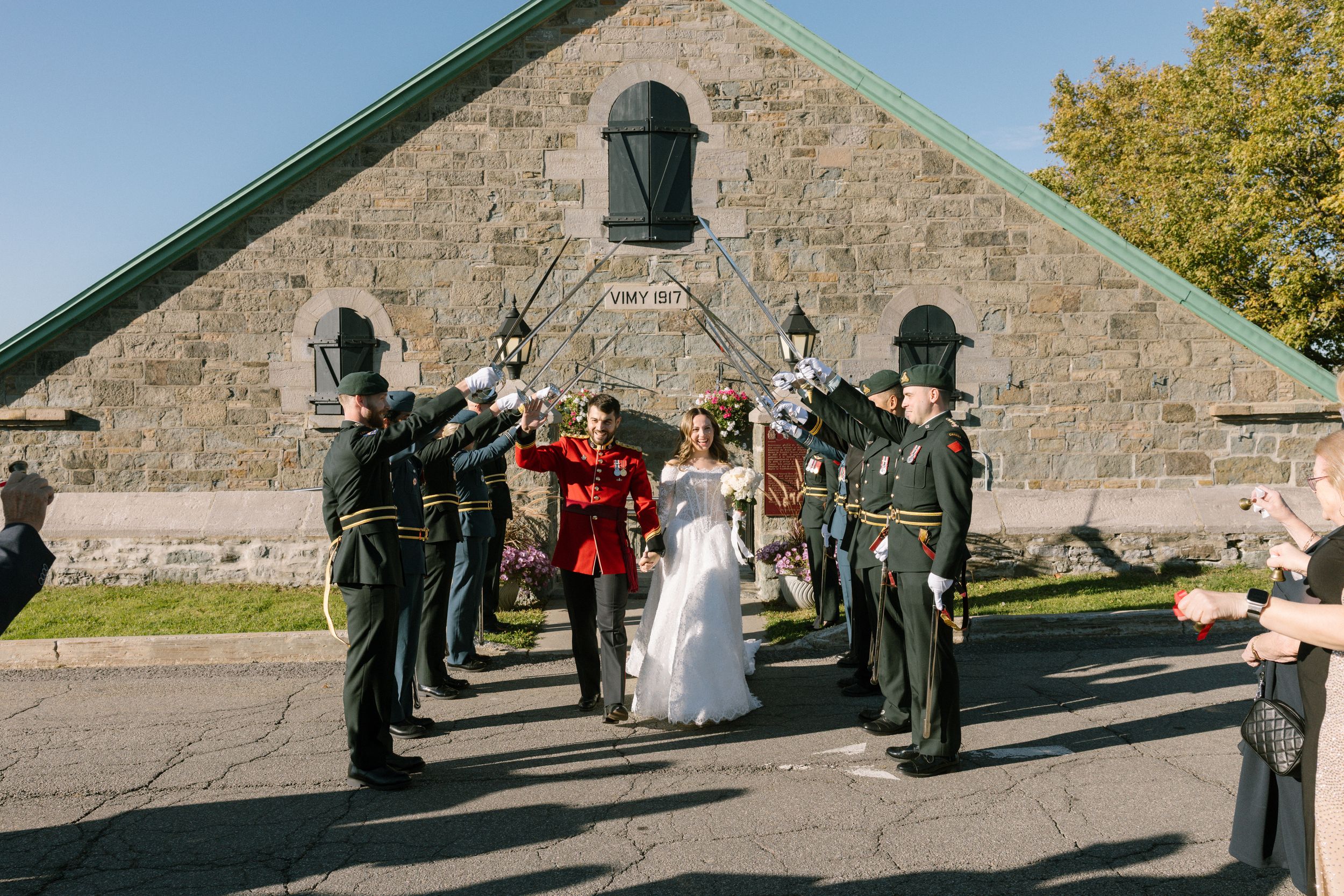 Military wedding ceremony with bride and groom walking under sword arch outside historic stone building.