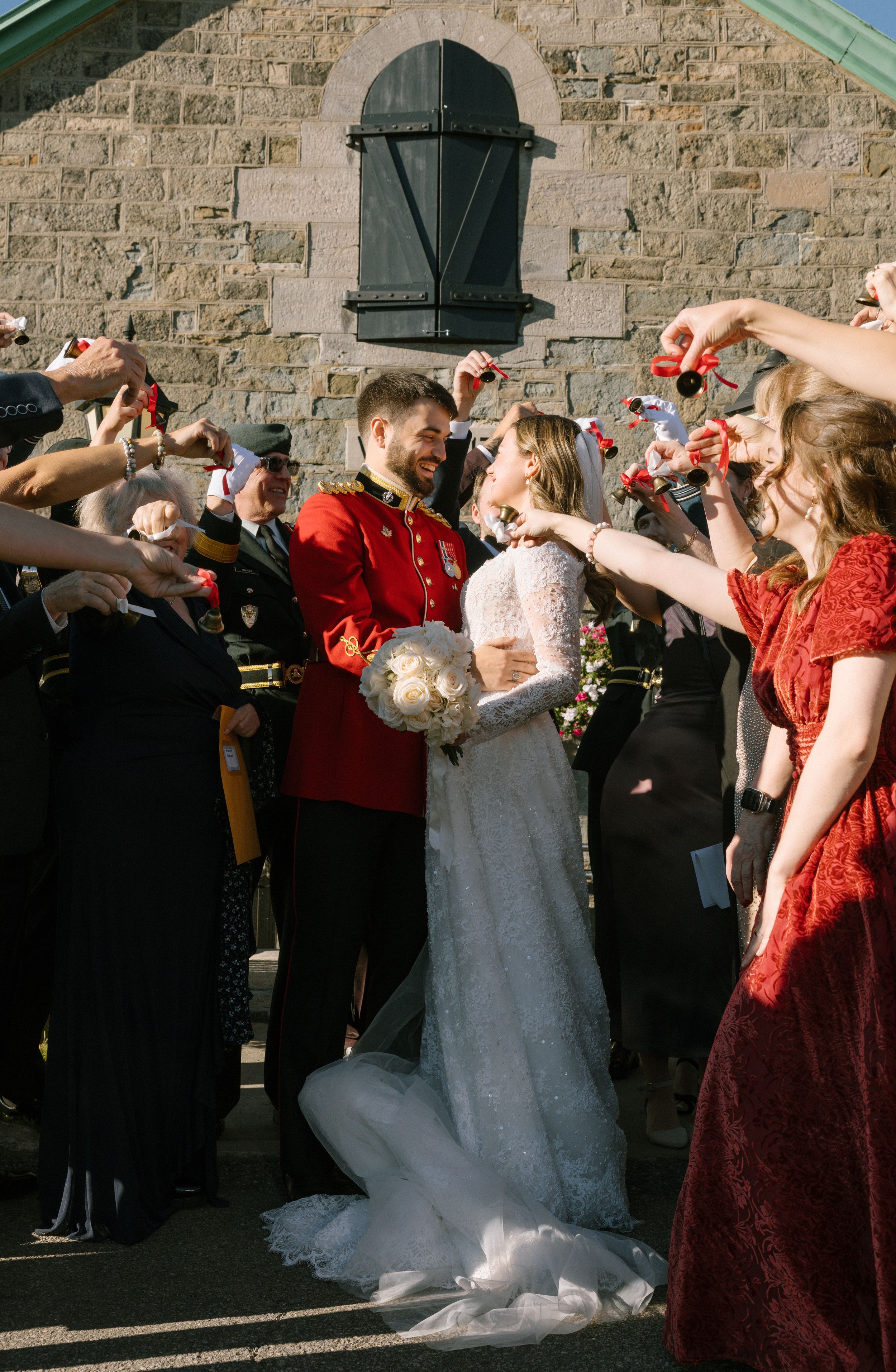 Military groom in red uniform kisses bride in white lace gown as wedding guests celebrate outside stone church.