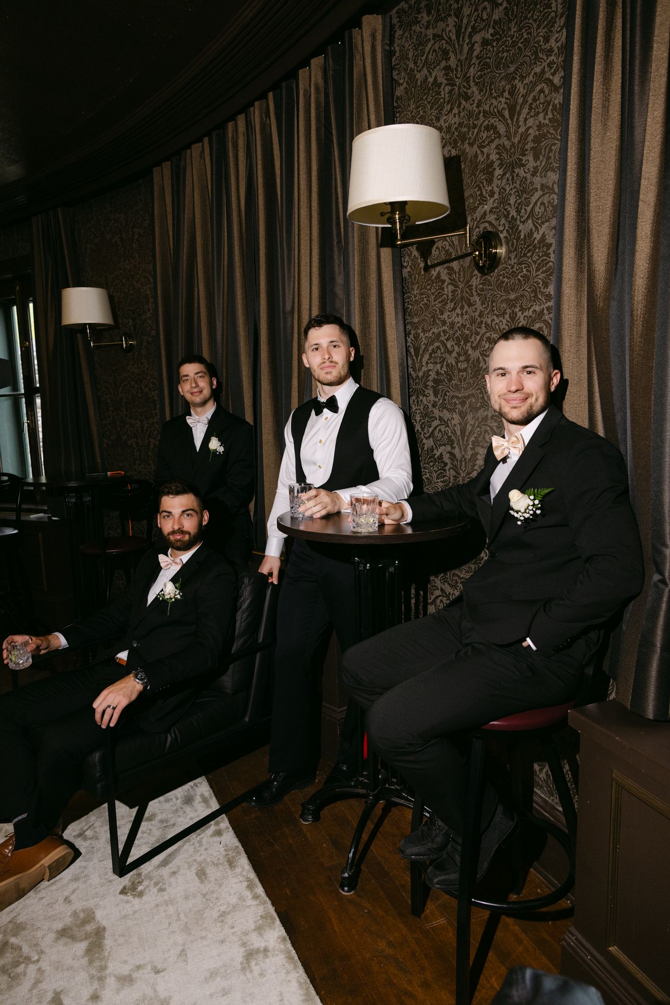 Four groomsmen in black suits with boutonnieres pose in an elegant dark-paneled room with draped curtains.