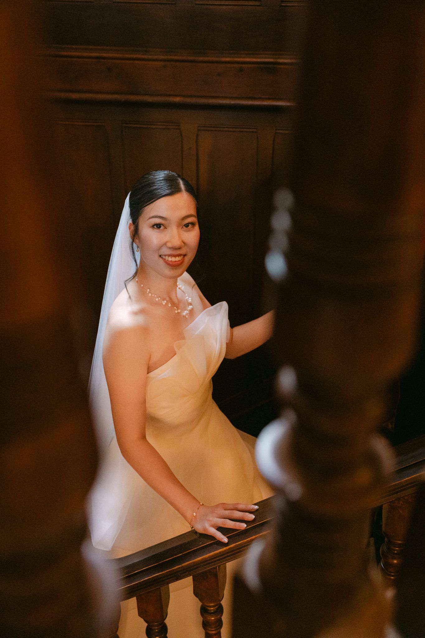 A smiling bride in a champagne strapless gown and veil poses gracefully on a wooden staircase.