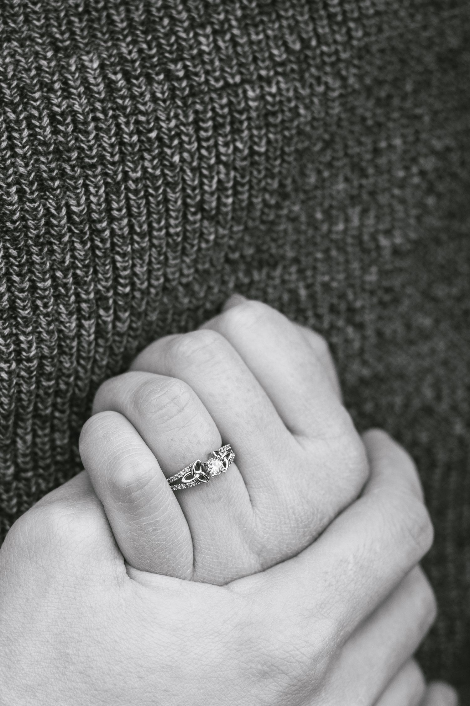 A delicate engagement ring sparkles on a hand against a textured gray knit background.
