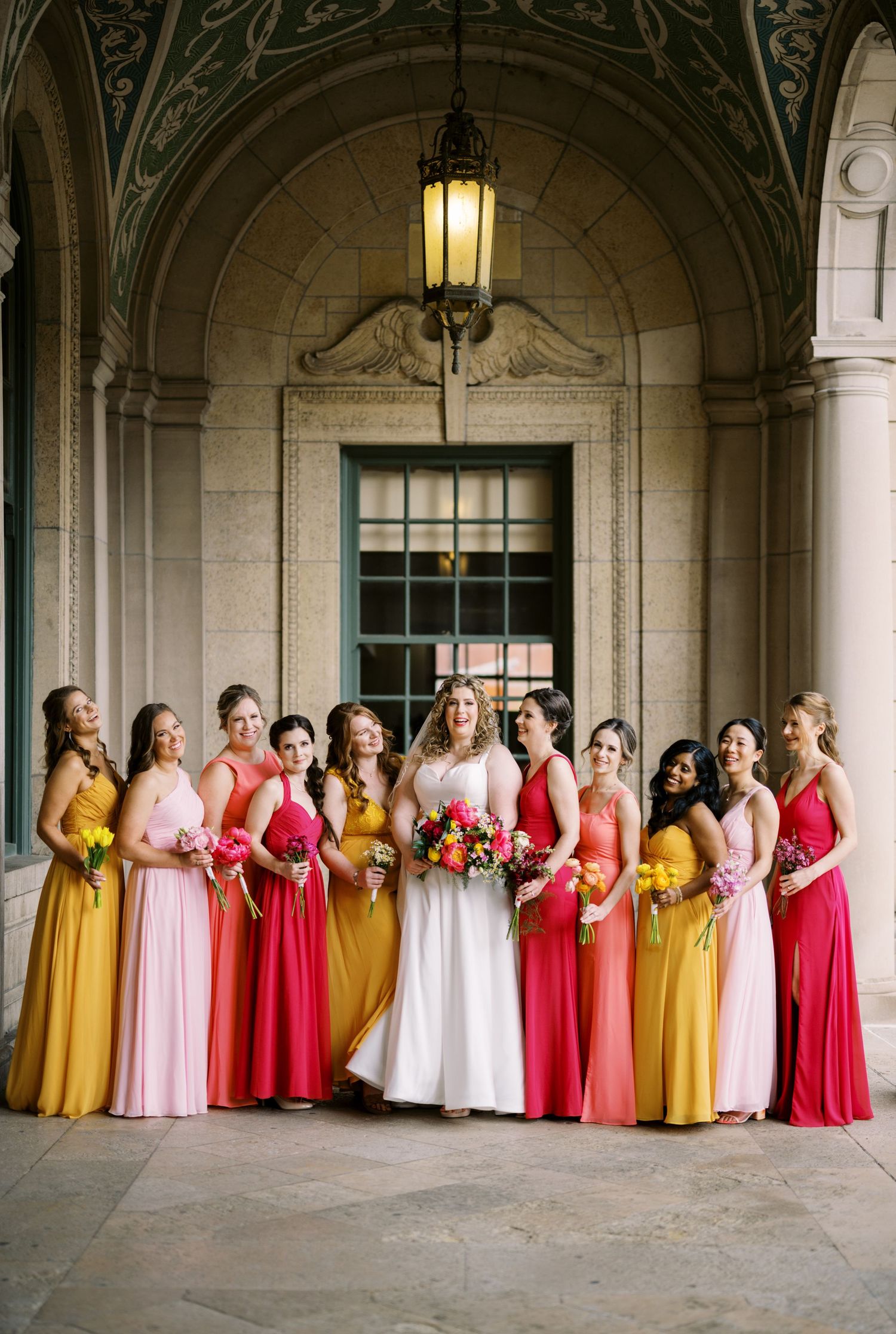 Wedding party in coral, yellow and pink flowing dresses posed under ornate stone archway.