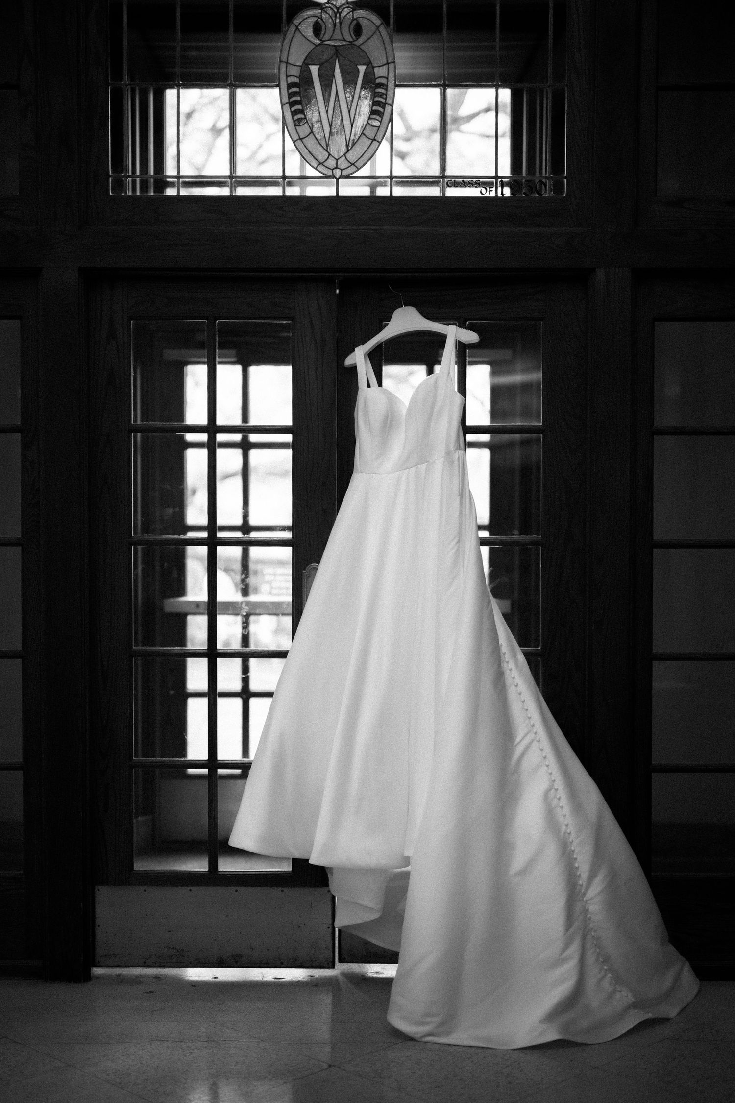 An elegant white wedding dress hanging in a dark window frame with ornate architectural details.