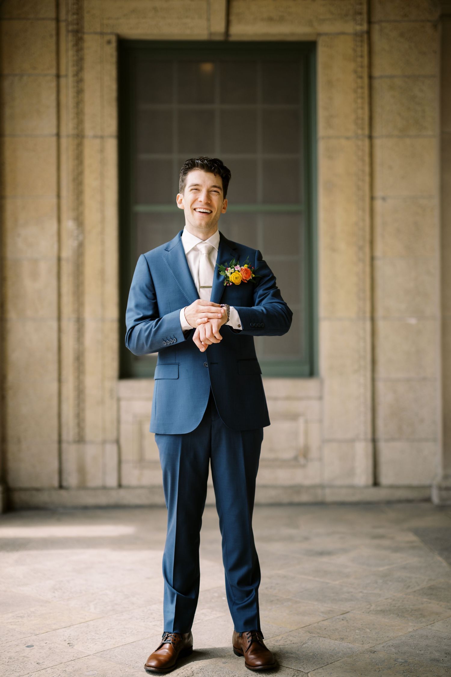 Navy blue suited figure with orange floral boutonniere standing in front of stone columns.