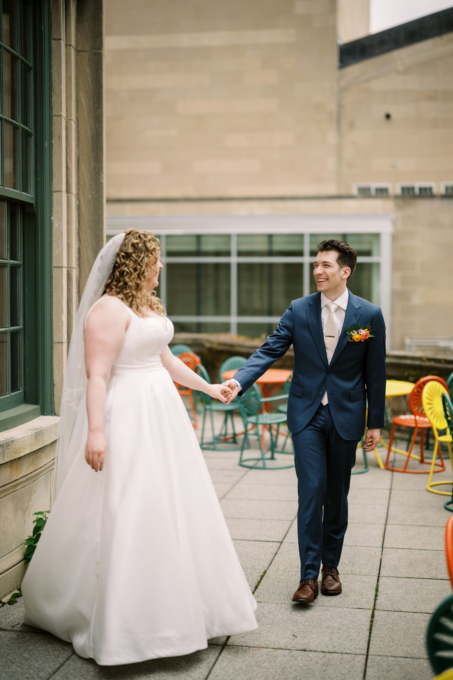 Wedding couple holding hands while walking on stone terrace with colorful chairs in background.