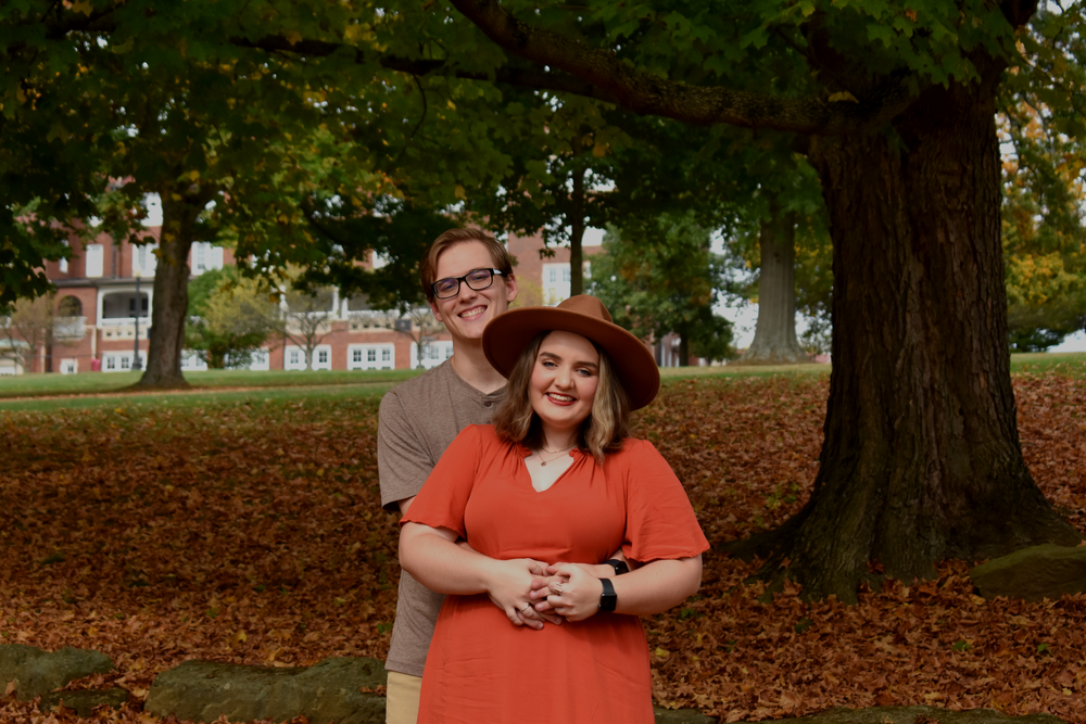 A couple stands beneath a large tree with fallen autumn leaves scattered around them.