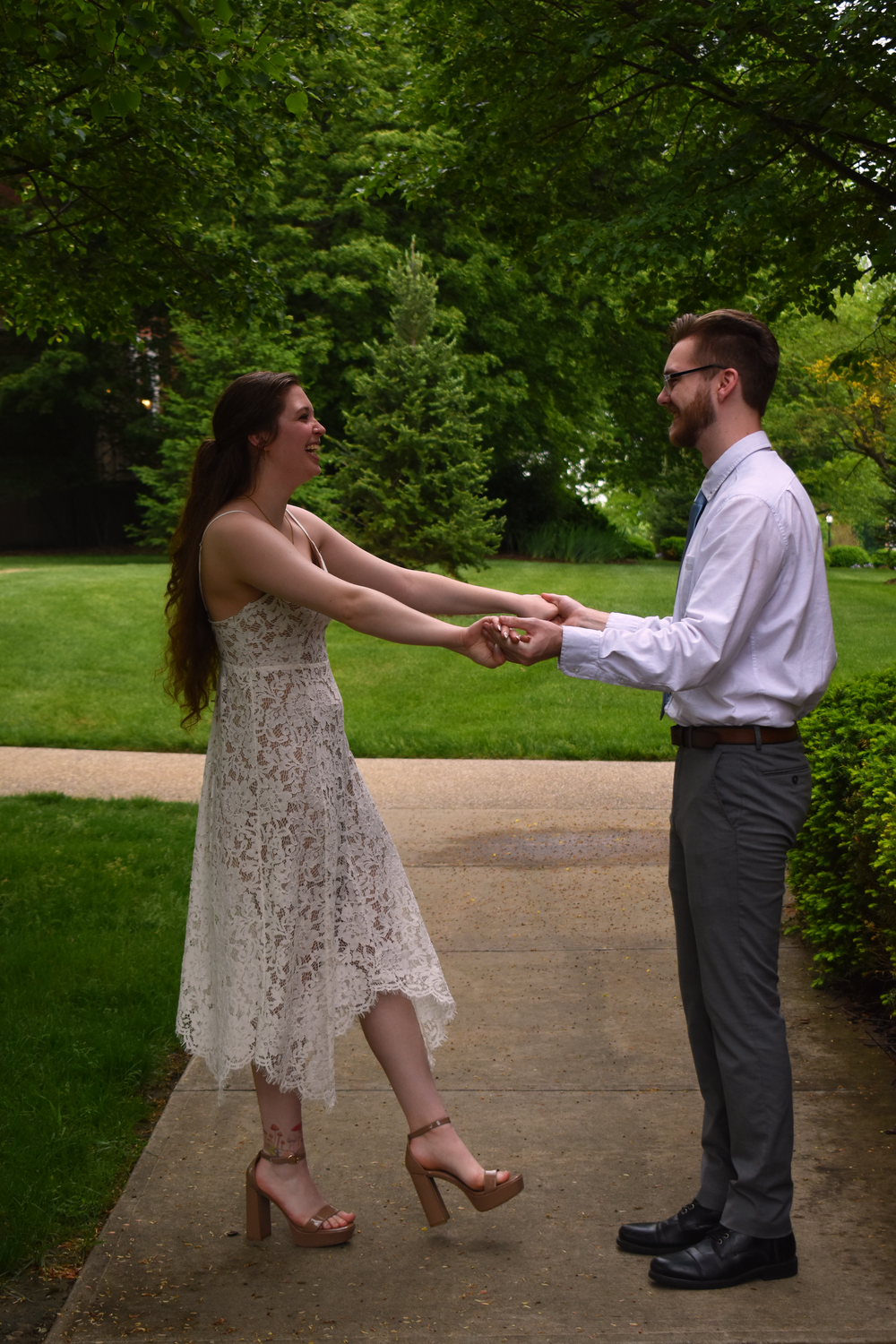 A couple in formal attire dancing together on a path surrounded by green trees.