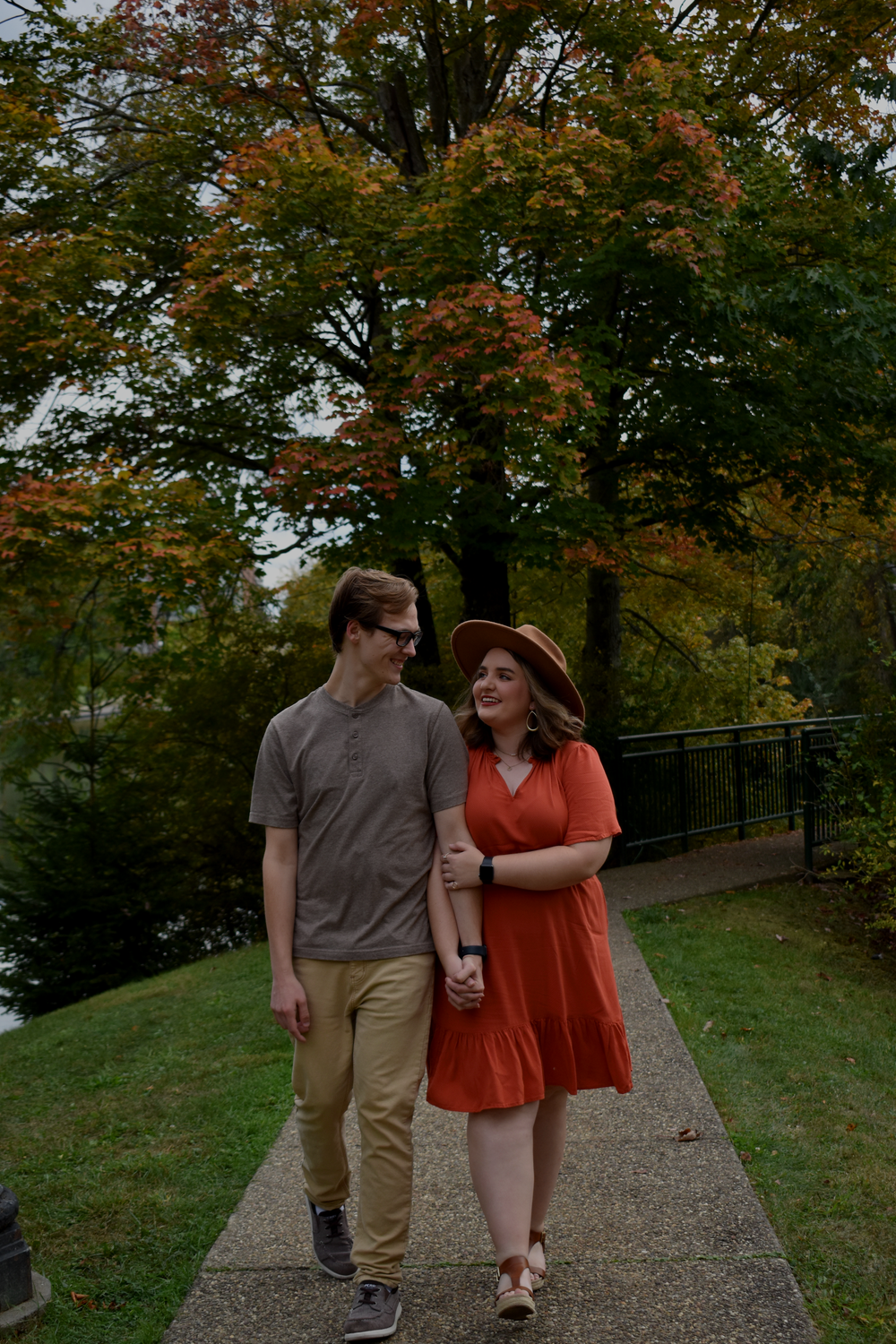 A couple walks hand in hand along a park path surrounded by autumn trees.