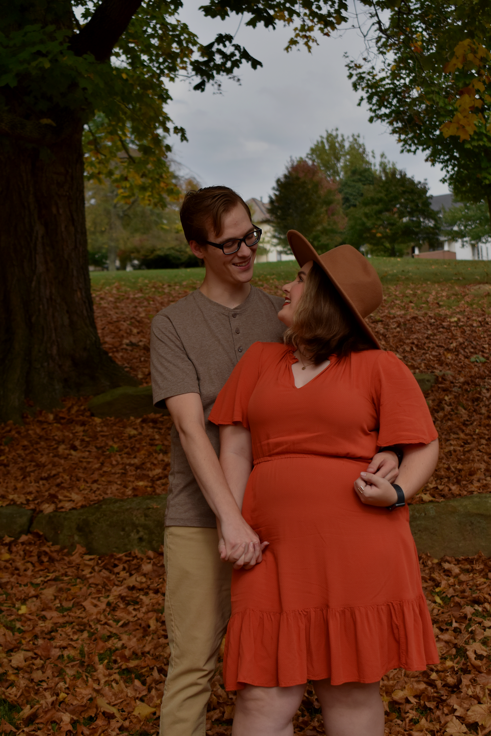 A couple in autumn attire poses affectionately among fallen leaves in a park setting.