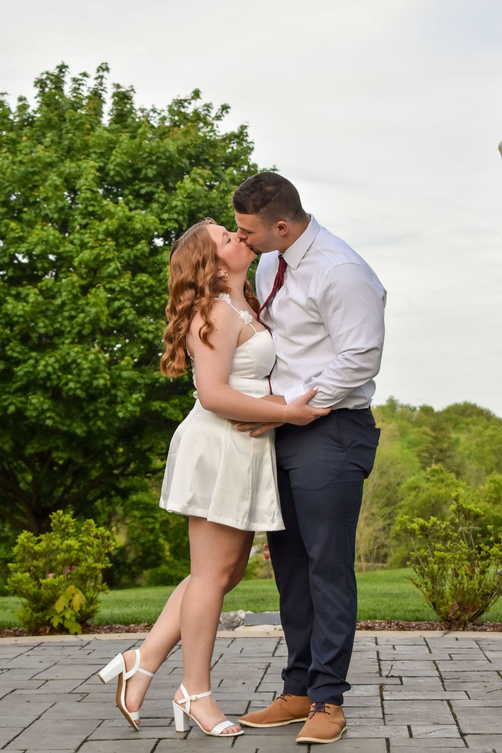 Two people share a romantic kiss while standing on stone patio with lush greenery in the background.