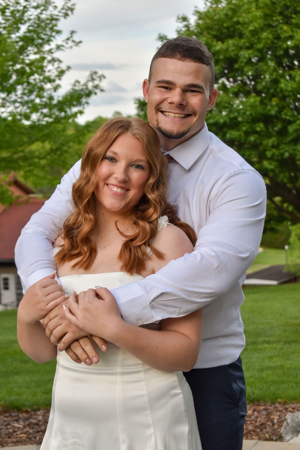 A couple embraces outdoors in formal attire with green trees and landscaping in the background.