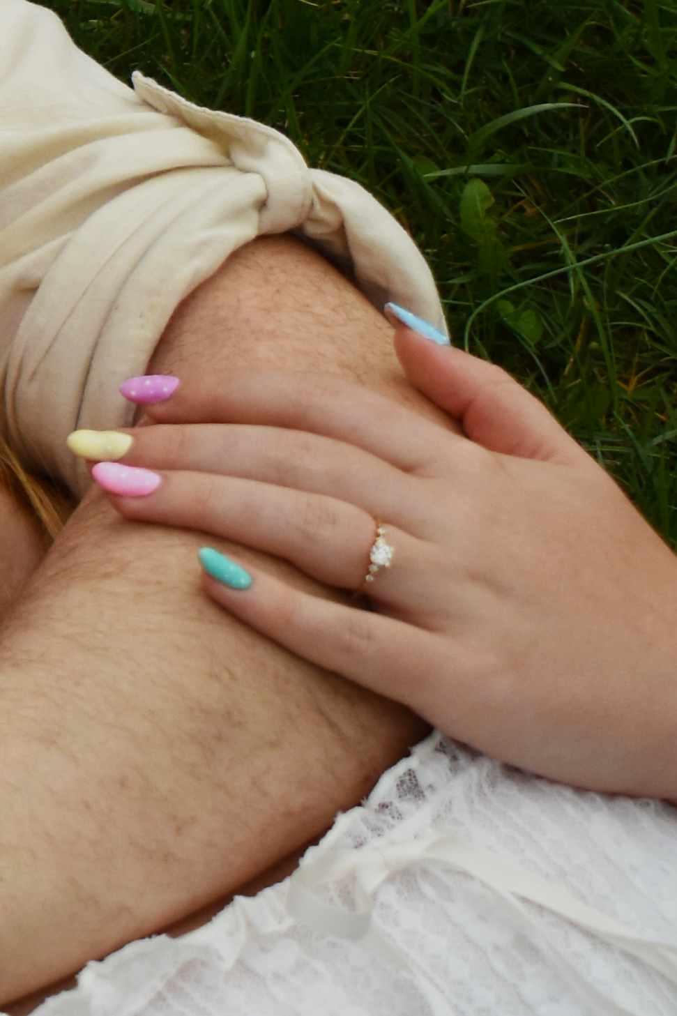 A hand with pastel pink and blue nails and a ring rests on white and cream fabric against green grass.
