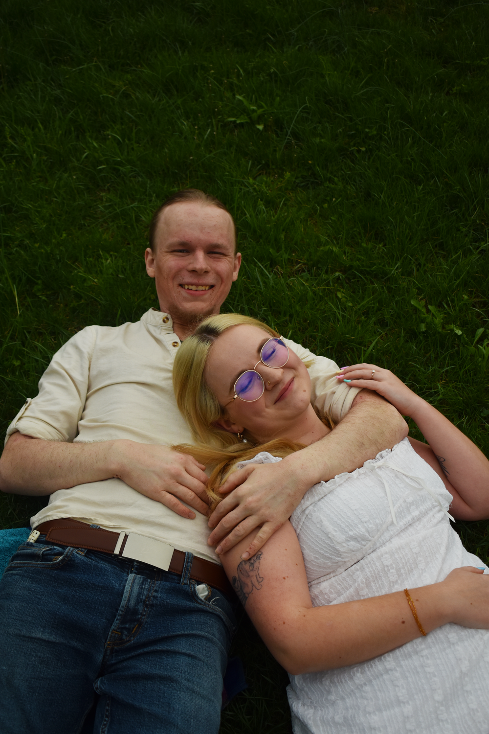 A couple relaxes together on green grass, with one person wearing a white dress and glasses leaning on their partner.