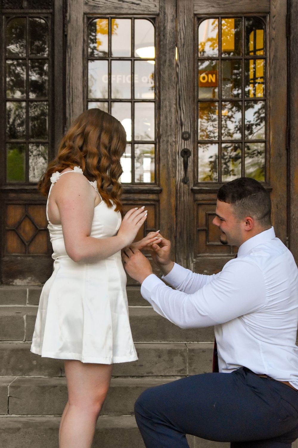 A romantic proposal takes place on stone steps in front of an ornate wooden doorway with glowing windows at sunset.