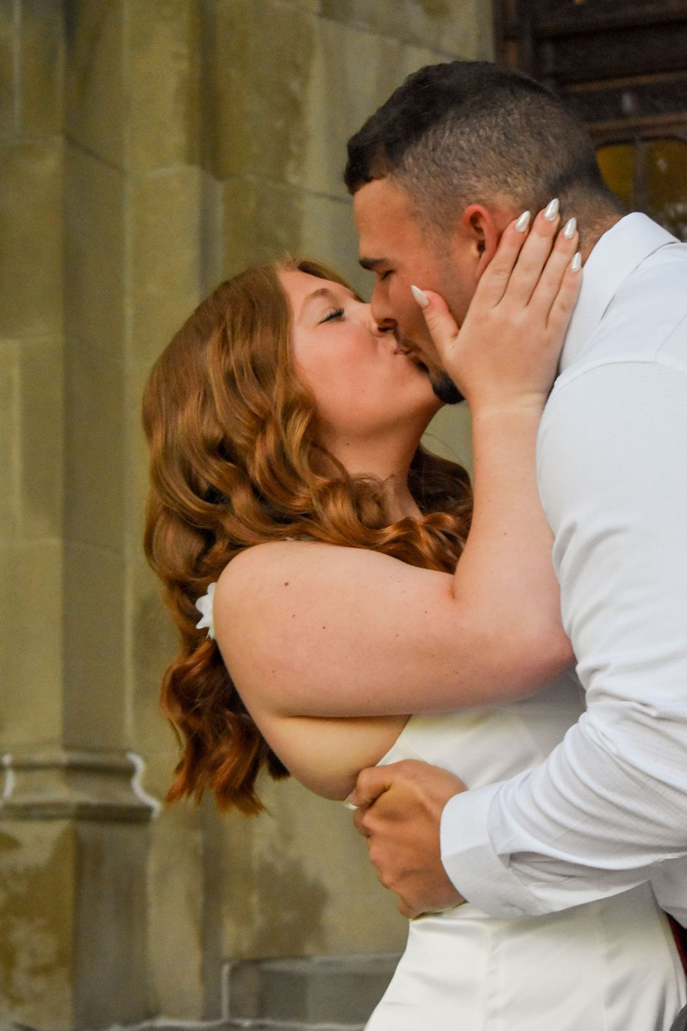 A romantic couple in white attire share an intimate kiss against a stone building wall.