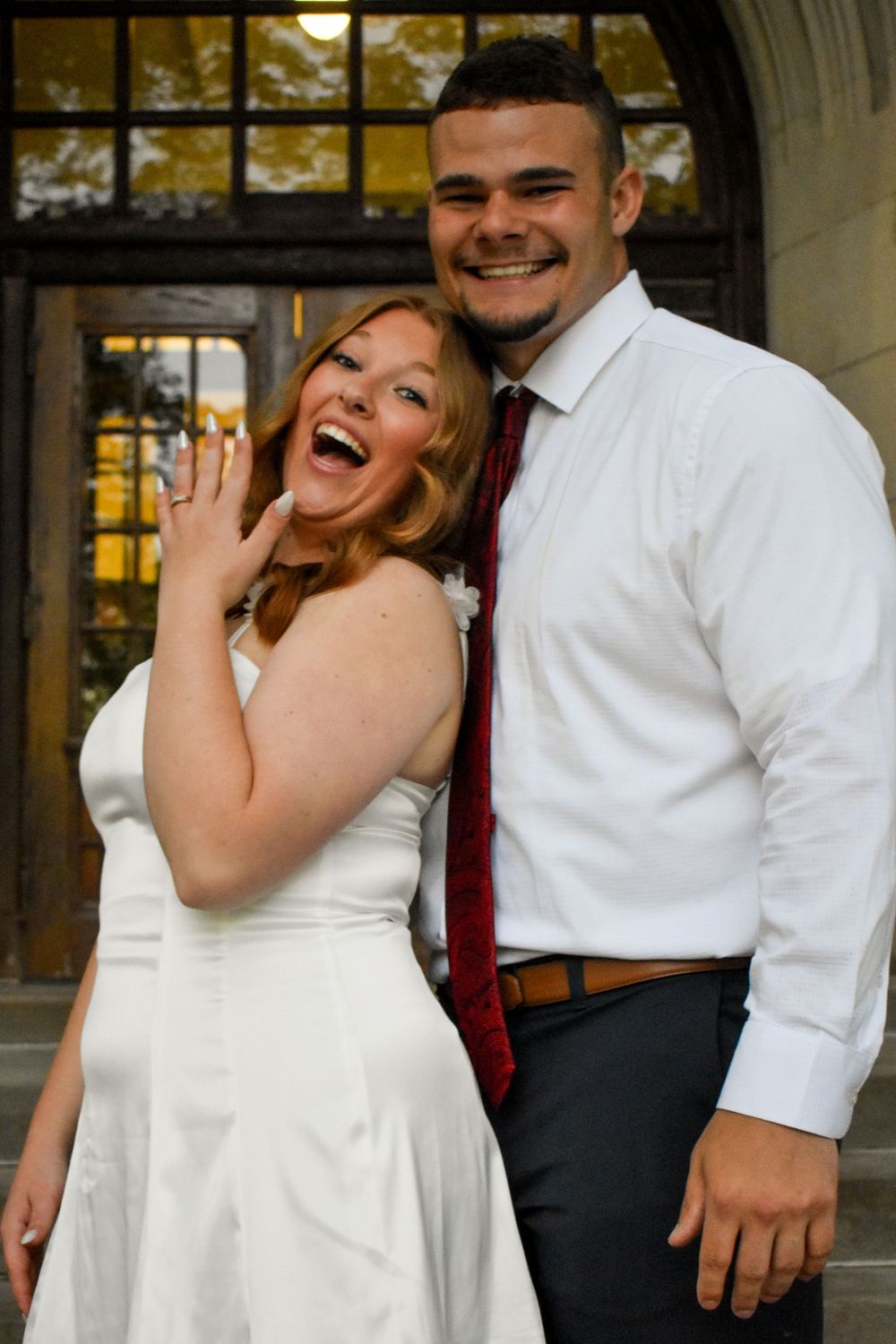 A joyful couple in formal attire smiles and poses together against an architectural backdrop with autumn colors visible.