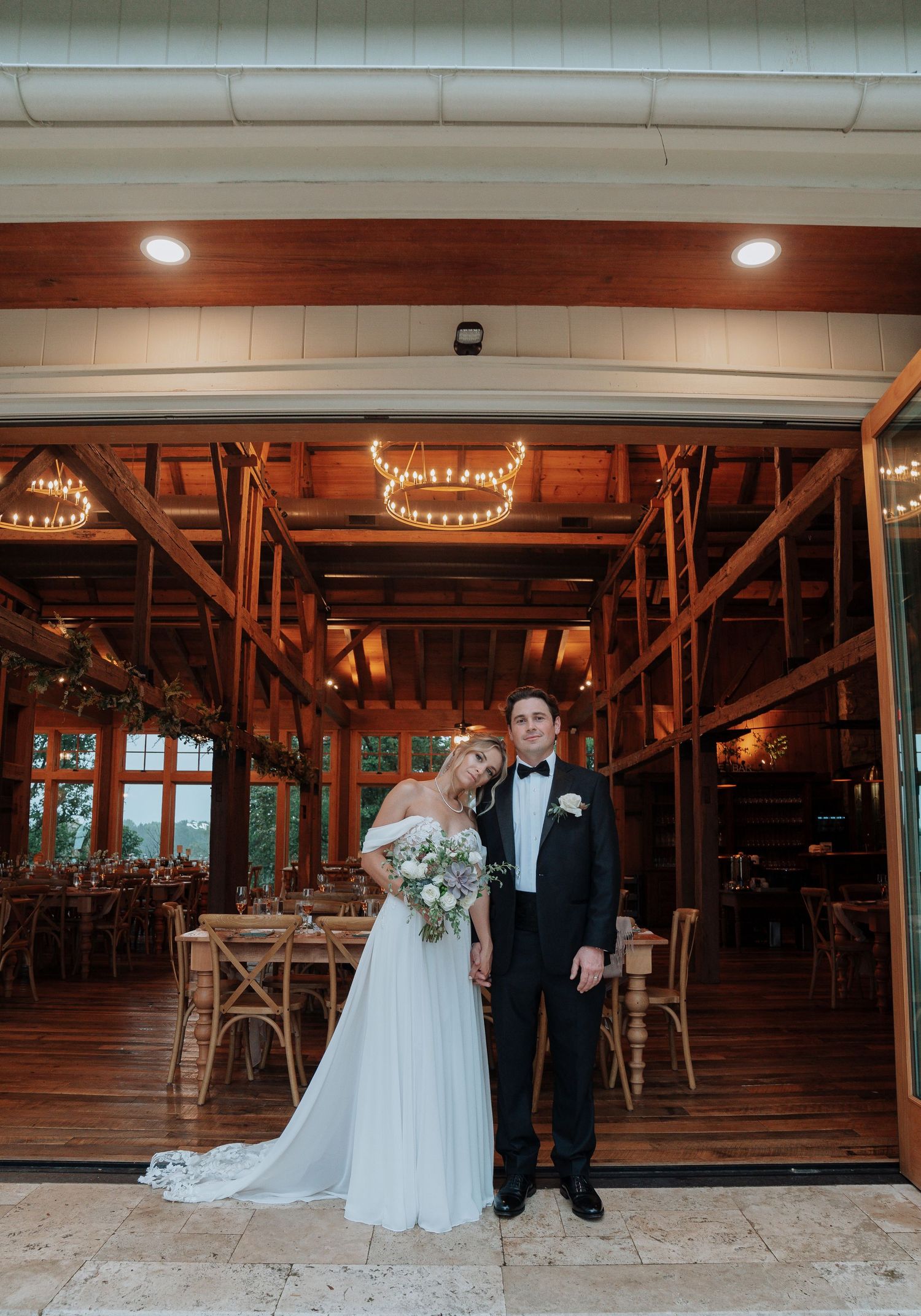 Elegant bride and groom pose together inside a warmly lit rustic barn wedding venue with string lights.