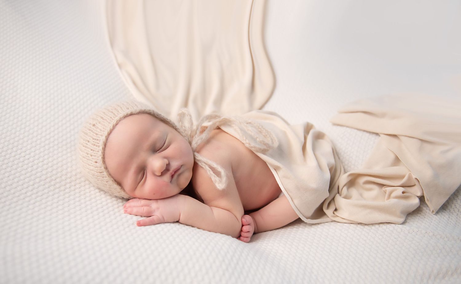 Peaceful sleeping newborn in cream wrap and bonnet on textured white backdrop.