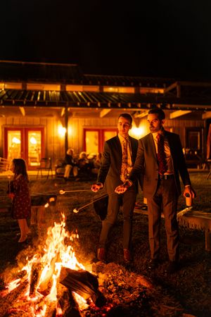 People gather around a glowing bonfire at night outside a rustic building with warm lighting.