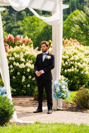 Groom in black suit with blue bow tie stands under white wedding arch surrounded by blooming gardens.
