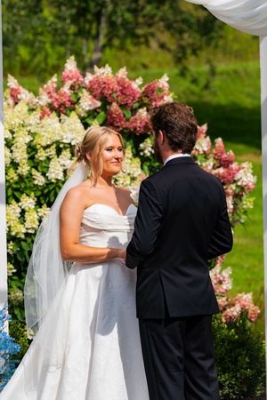 Bride in white strapless gown holds hands with groom at outdoor wedding ceremony with pink hydrangeas.