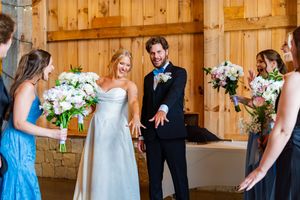 Smiling bride and groom walk down aisle in rustic barn venue, surrounded by bridesmaids holding pink and white bouquets.