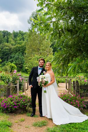 Bride and groom pose in lush garden with pink flowers, bride holding bouquet in white gown, groom in black tuxedo.