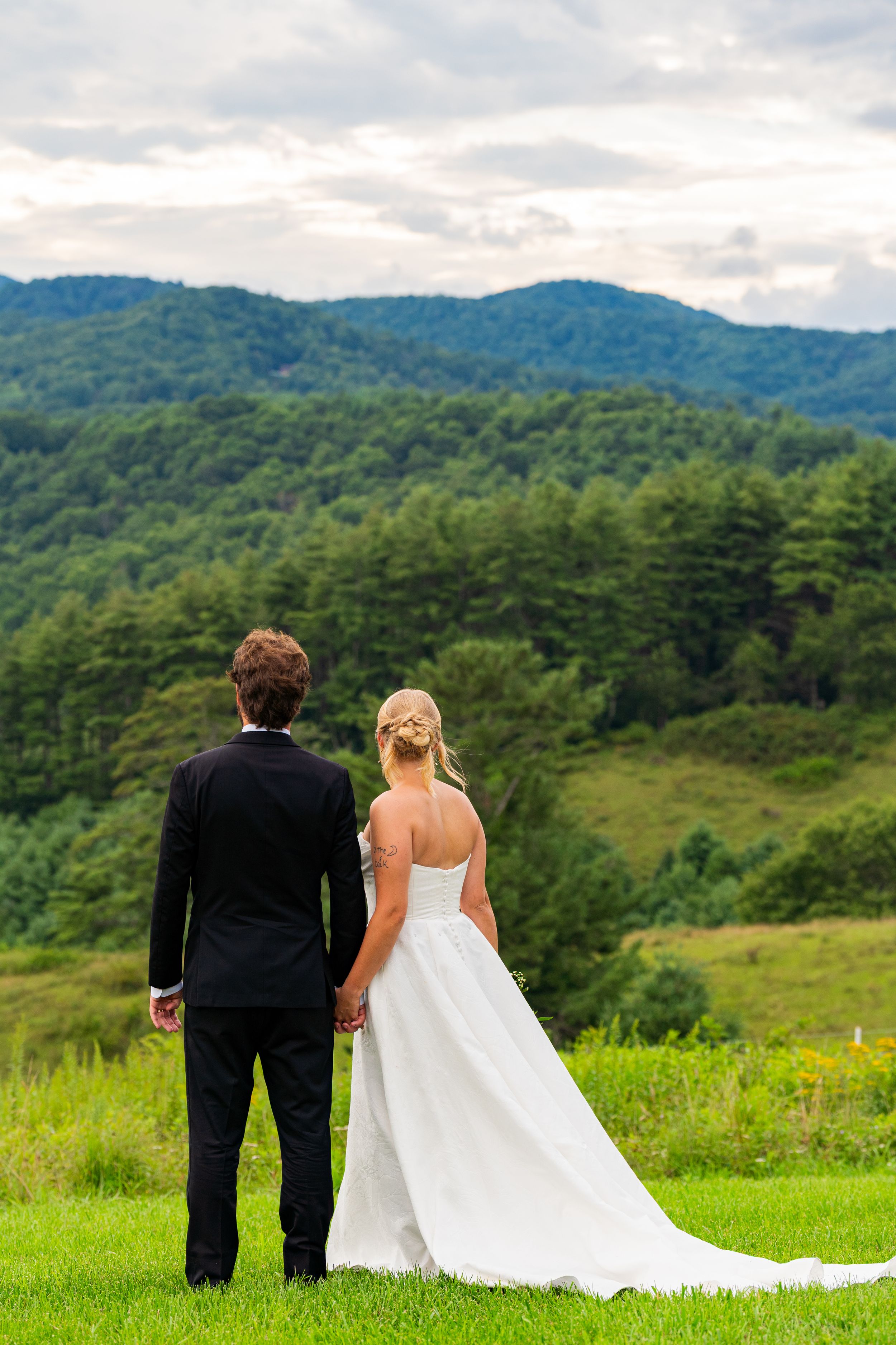 Bride and groom holding hands facing lush green mountains on their wedding day.