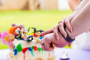 Couple cutting a colorful wedding cake together outdoors, hands joined with rings visible.