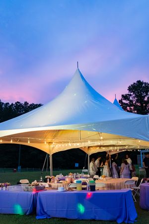 White event tent glowing with string lights at dusk, hosting an outdoor party with purple-lit tables under a colorful sunset sky.