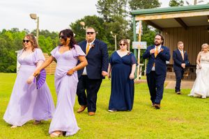 Wedding party walks outdoors in lavender and navy formal attire on green lawn near rustic pavilion.