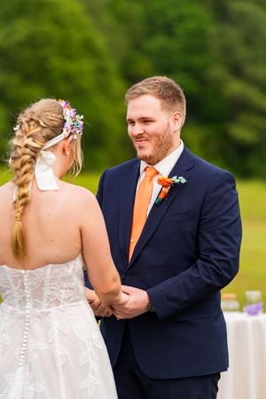Groom in navy suit with orange tie holds bride's hands during outdoor wedding ceremony.