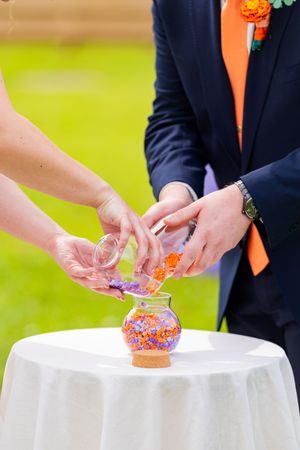 Bride and groom pouring colorful sand into a glass vase during a unity sand ceremony at an outdoor wedding.