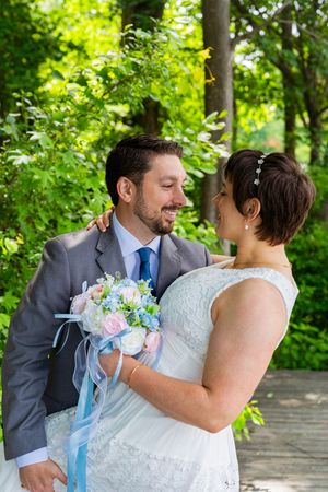 Bride and groom smiling at each other outdoors, holding a pink and blue bouquet with ribbon, surrounded by lush green trees.