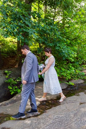 Bride in white dress holding bouquet and groom in gray suit walk hand in hand on rocky path in lush green park.
