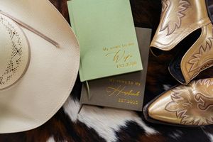 Western wedding flat lay with cowboy hat, gold boots, and personalized vow books on cowhide.