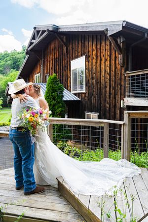 Bride in white gown with bouquet embraces groom in cowboy hat on rustic wooden deck outside barn venue.